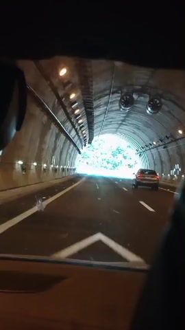 The video captures a first-person perspective from inside a vehicle as it exits a tunnel. The tunnel interior is visible with lights lining the ceiling and ribbed walls. As the vehicle emerges, daylight reveals a paved road with white lane markings. A car is seen ahead in the same lane. The exit leads to an elevated roadway with a mountainous, tree-covered landscape in the background. Traffic signs are visible on the side of the road, including a yellow sign with a pictogram of a person working with a shovel and a blue sign with a white symbol. The video ends as the vehicle continues on the open road.