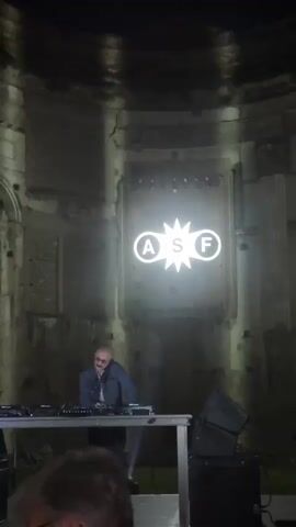 A bald man with glasses stands behind a DJ booth, operating the equipment. The booth is positioned in front of a large, dilapidated structure with a curved ceiling. A bright white logo with the letters "ASF" inside circles and a starburst is projected onto the wall behind the DJ. The scene appears to be an outdoor event at night.