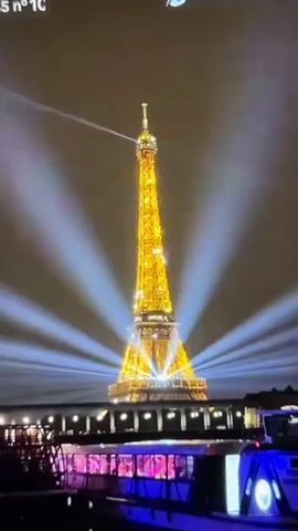 The video shows the Eiffel Tower illuminated at night with golden lights and a display of blue spotlights radiating from its base. The tower stands tall against a dark, hazy sky. In the foreground, a boat with illuminated windows is partially visible, suggesting the scene is viewed from the water, likely the Seine River. The overall atmosphere is festive and grand.