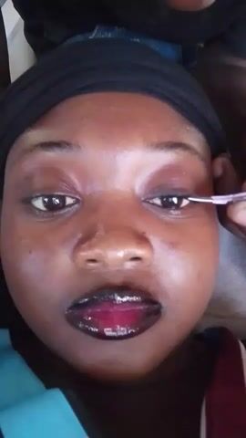 A close-up shot shows a person with dark skin, wearing a black headwrap, looking directly at the camera. Their lips are coated in dark lipstick with a reddish-purple center. A second person's hand, holding pink tweezers, is visible in the upper right of the frame, near the subject's right eye. The tweezers appear to be manipulating something around the subject's eyelashes or eyebrow area. The background is out of focus.