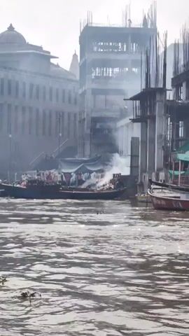 A wide shot captures a riverfront scene dominated by a large building under construction, its scaffolding and unfinished concrete structure stark against a hazy sky. In the foreground, the murky river flows, dotted with green aquatic plants. A long boat, laden with goods and several people, is moored near the shore, with smoke rising from it, suggesting some activity like cooking or burning. Another smaller boat is tied nearby. The backdrop features more traditional-looking buildings, one with a dome-like structure. The overall atmosphere is one of a busy, working waterfront, with a contrast between old architecture and modern development.