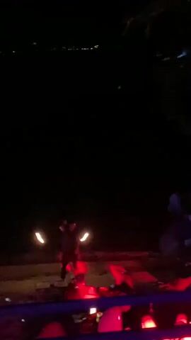 The video shows a dark outdoor scene at night, likely a restaurant or lounge. In the distance, a person is performing with two lit objects that appear to be on fire. Close by, there are tables and chairs illuminated with red lighting. Several people are seated and socializing. The foreground includes leaves from an overhead tree. The scene appears to be set in a tropical environment, as indicated by the lush vegetation.