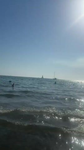 A bright sunny day at the beach. The camera is angled towards the ocean, capturing the water and the sky. Gentle waves are rolling in, with sunlight glinting off the surface. In the distance, several sailboats are visible on the horizon. There are a few people in the water, swimming or wading. The water appears clear, and the sky is a brilliant blue. The overall atmosphere is peaceful and idyllic.