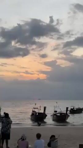 A beach scene at sunset shows several longtail boats in the water. Three people sit on the beach while another person, possibly taking photos or videos, stands. The sky is cloudy with a glimpse of the setting sun on the horizon. The water is calm and the boats are gently bobbing.