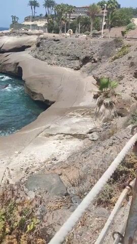 The video shows a rocky coastline with the ocean on the left. Waves are crashing against the shore. In the distance, a small boat with two masts is visible on the water. The sky is clear and blue. The vegetation on the shore is dry and sparse, consisting of grasses and small bushes. The terrain is uneven and rocky. The scene appears to be a natural landscape.