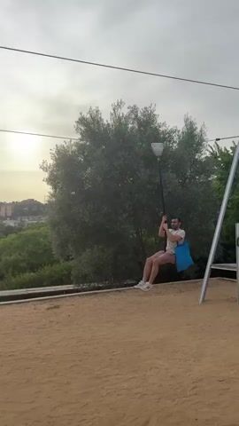 A man on a zipline moves from left to right across the frame. He is holding onto a pole with both hands and has a blue bag hanging from his left side. He is wearing shorts and a t-shirt. The zipline is suspended between two poles, and the man is moving over a sandy area. In the background, there are trees, buildings, and a hill under a cloudy sky. The sun is visible in the upper left corner of the frame.