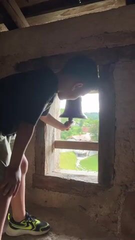 A young boy, seen from the side, is ringing a bell out of a window of a stone building. He is wearing a dark t-shirt, light-colored shorts, and athletic shoes. The bell is dark and held by a rope. Through the window, a green, hilly landscape with houses can be seen. The interior of the building appears to be made of stone and wood.