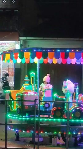 Here's a description of the video:

A small, illuminated carousel is running in front of a storefront at night. The carousel is decorated with colorful lights outlining its base and canopy, which is topped with multicolored "scallops." It features various ride-on vehicles, including a green and yellow toy train and a white Hello Kitty figure. A child wearing a light-colored hat is seated in the train, appearing to enjoy the ride.

In the background, you can see a store with a sign that says "AH AGENCY" and some visible products inside. There is a scooter parked in front of the store. The overall lighting is dim, with the carousel lights providing the main source of illumination.