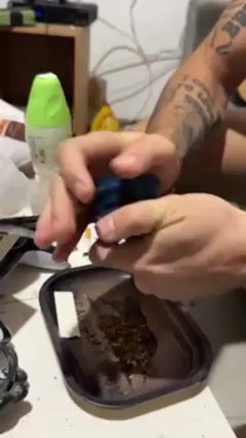 The video shows a person sitting at a table, grinding something in a blue grinder. The person is using their hands to twist the grinder over a black rolling tray containing brown plant matter and a white filter tip. The person has tattoos on their arm and is wearing a gold bracelet. Other objects on the table include a green air freshener, a bag of chips, a pair of headphones, and a watch. In the background, there is a wooden shelf with electronic devices on it. The lighting appears to be artificial.