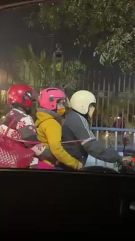The video shows a family of three riding a motorcycle at night. The driver is wearing a white helmet and a gray jacket. The passenger behind him is wearing a pink helmet and a yellow jacket. The passenger behind them is wearing a red helmet and a printed jacket, and holding a large red bag. They are stopped at a traffic light. The view is from inside a car. Another motorcycle passes by.