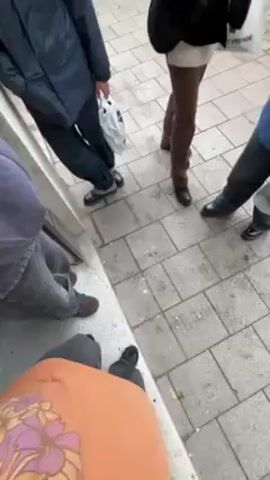 The video shows a person opening a door to a clothing store. Outside, a group of people are standing on a tiled sidewalk. One person is holding a white plastic bag. Inside the store, there are racks of clothing, including colorful t-shirts. There are also large plastic bags filled with clothes, and a staircase leading to a loft area. The store appears somewhat cluttered.
