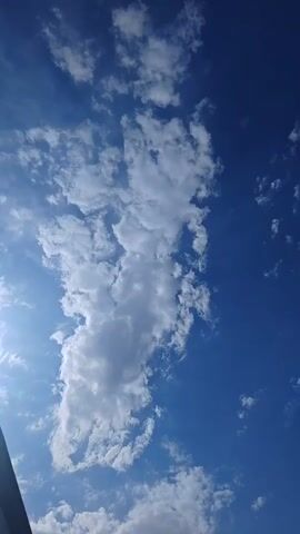 The video displays a blue sky with scattered white clouds. The camera appears to be tilted upwards, capturing the sky from a low angle. Sunlight is visible, suggesting it is daytime. The clouds are wispy and varied in size, with some appearing more dense and others more translucent. There is no indication of people, objects, or any specific actions taking place within the frame. The environment is solely the sky and clouds.