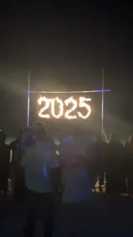 A crowd of people, mostly silhouetted, are gathered at night, facing a large sign that reads "2025" in burning flames. The sign is supported by a metal frame. The overall environment appears dark, with light emanating from the sign. Some individuals are holding up their phones.