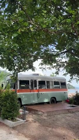 A stationary vintage bus, painted in light green with an orange stripe, is parked on a paved area next to a dirt path under the shade of a large tree. The bus has multiple windows and appears to be unoccupied. In the background, beyond a grassy area, is a body of water with a rocky shoreline. A small red object, possibly a kayak or raft, is visible near the water. The sky is partly cloudy. The video pans slowly to the right, revealing more of the tree and the background scenery.