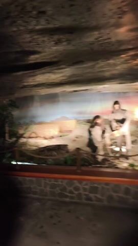 The video shows a diorama recreating a Neolithic salt works scene, likely in a museum or historical exhibit. The diorama contains life-sized figures dressed in primitive clothing, engaged in activities related to salt extraction, such as tending a fire and working with tools. The backdrop consists of a painted landscape, while natural elements like straw and plants are used for added realism. A nearby plaque reads "Neolityczna Warzelnia Soli Sprzed 6 Tys. Lat," which translates to "Neolithic Saline Works 6 Thousand Years Ago." The room is dimly lit, with the ceiling appearing to be the uneven surface of a cave. The area in front of the diorama has a floor that resembles stone tiling.