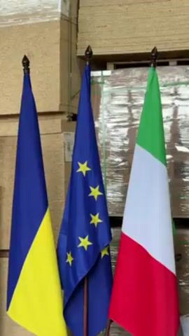 The video shows three flags standing in front of a backdrop of stacked boxes. From left to right, the flags are the flag of Ukraine (blue over yellow), the flag of the European Union (a blue field with twelve yellow stars), and the flag of Italy (green, white, and red vertical stripes). All three flags have decorative finials at the top of their flagpoles.