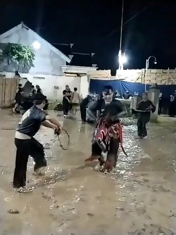 At night, an outdoor cultural performance takes place on very muddy ground, likely in Jenggrik, Indonesia. Numerous individuals are present, with artificial lights illuminating the scene. In the foreground, a man in a black shirt and pants with a design on his back is actively dancing or performing, holding a prop that resembles a short pole or stick. Another individual, dressed in an ornate red and black *kuda lumping* (hobby horse) costume, moves alongside him, mimicking the movements of a horse.

The ground is saturated with mud and standing water, suggesting recent rain or that it is an integral part of the performance setting. In the background, other people observe the activity or are gathered around. Structures like a white building, a wooden fence-like barrier, and a blue tarp are visible. Tall poles with flags are also seen in the distance, further indicating a community event or celebration. The specific design on the black shirt and the horse costume are visible but not clear enough to read any text.