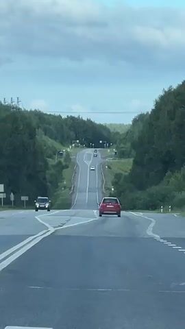 A camera captures a view from a car driving on a highway surrounded by trees. The road stretches into the distance, with a series of hills and curves. Several other cars are visible on the road, some ahead and some approaching. The sky is cloudy with patches of blue. The car's dashboard and steering wheel are visible in the foreground of the later frames. The overall impression is of a scenic drive through a rural landscape.