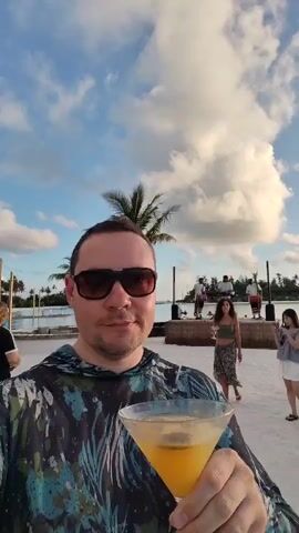A man in sunglasses and a patterned hoodie smiles at the camera while holding a martini glass with an orange drink. He is standing on a sandy beach with palm trees and a body of water in the background. In the distance, a band performs on a stage, and several other people are visible, including a woman in a green top and a young girl in a white dress. The sky is blue with fluffy white clouds, and the lighting suggests it is late afternoon or early evening.
