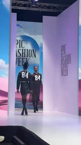 A fashion show is in progress. The backdrop features the text “Epic Fashion Week.” Two female models are walking down the runway, wearing outfits in shades of black. One model is in a short black leather dress and knee-high socks, while the other wears a black mesh dress. Both outfits are numbered 84 in white. The model in the black mesh dress is in the foreground, while the other model is behind her. The text on the screen transitions from “Epic Fashion Week” to “Achirima Lagos.” The set is framed by large white panels, and the runway is brightly lit.