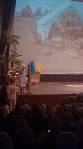 A stage performance in front of a seated audience shows actors on a platform with props including palm trees, a camel with lights, and colorful cloth, set against a backdrop of a desert camp scene projected on a screen. The lighting is dim, and the video captures audience members watching.