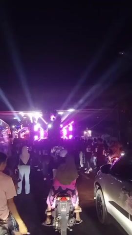 The video shows a night scene of a crowded outdoor event, possibly a concert or festival. A stage is set up with bright pink and white lights, beams of light projecting upwards into the dark sky. Several people are gathered in front of the stage, with many more visible further back. A motorcycle is parked in the foreground, with a person seated on it facing away from the camera. Another person is partially visible in the lower left corner of the frame. A silver car is parked on the right side of the road. The overall atmosphere appears to be lively and energetic.