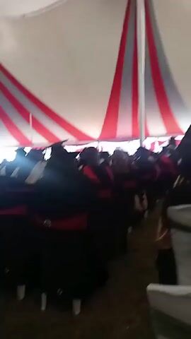 The video shows a graduation ceremony taking place inside a large tent. The tent has a white ceiling with alternating red and white stripes running down the sides.  Many people, presumably graduates, are seated in rows of white chairs. The graduates are wearing black graduation gowns and caps. Some of them have red stoles, while others have gold or tan stoles. The camera is positioned behind the seated graduates, facing towards the front of the tent.