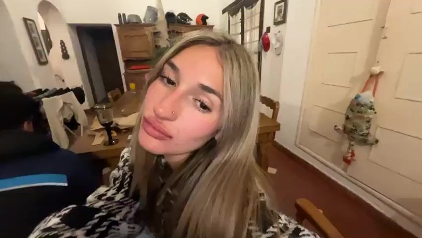 The video shows a close-up of a woman with long blonde hair, wearing a black and white patterned jacket. She is in what appears to be a dimly lit room with furniture and objects visible in the background, including a wooden table with items on it, a chair, and what looks like a door. The woman's eyes are mostly closed, and she appears to be very tired or possibly falling asleep. There is a piercing on her nose. A portion of another person's shoulder is visible in the lower left corner of the frame.