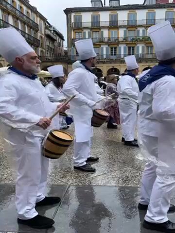 Okay, here's a concise description of the video:

The video shows a group of people dressed in matching white outfits and tall white hats, with some wearing blue neck scarves. They are performing in a public square, each playing a small drum with a stick. The square appears to be paved with light gray tiles, and there are buildings surrounding the square on all sides with multiple windows and balconies. The weather appears overcast. The people are moving in a circular formation as they play their drums. Several spectators are in the crowd.