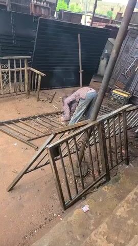 A man in a plaid shirt and light-colored pants is bent over, performing arc welding on a metal gate section lying on the reddish-brown dirt ground. Bright sparks emanate from the welding point. Surrounding him are several other unfinished metal gate or railing components, some lying flat and others propped up. In the background, large dark metal panels and ornate metal gates are stored upright. The scene appears to be an outdoor metal fabrication yard or workshop.