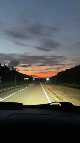 The video shows a first-person view from inside a car driving on a highway at dusk. The sky is a blend of dark blue and orange hues, with scattered clouds. The headlights of the car illuminate the asphalt road, which has white lane markings. Trees and utility poles line the sides of the highway. The sun is setting on the horizon, creating a bright orange glow. There are no other vehicles or people visible. The overall impression is of a peaceful drive through the countryside as daylight fades.