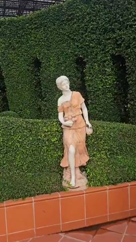 The video pans across a garden featuring meticulously trimmed hedges and terracotta tiled flooring. A statue of a woman in a flowing orange dress holding flowers stands near a large, potted bonsai tree with pink blossoms. Further in the background, a gazebo with ornate columns and a wrought-iron dome is visible, also surrounded by greenery and featuring another statue of a woman. A large pot with water lilies sits in the foreground. The scene appears to be an outdoor, landscaped garden.