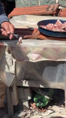 A close-up shot shows a person's hands using a fork to place pieces of raw meat on a flat black surface, possibly a griddle or cooking stone. The meat is red with white marbling, and some pieces appear to be pork belly. The cooking surface is placed over a makeshift fire pit constructed from bricks, with some smoldering embers visible beneath. In the background, a green plastic bottle is seen among scattered debris and wood. The scene appears to be outdoors, with a plastic tarp providing some cover overhead. The overall setting suggests an informal outdoor cooking arrangement.