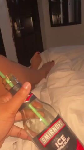 A hand holds a bottle of Smirnoff Ice Double Black with a green straw in it. The bottle is almost empty. The hand is resting on a white bed. Bare feet are visible in the background. A door and a framed picture are visible in the blurry background.