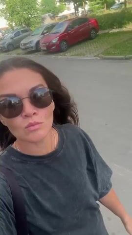 A woman with long dark hair, wearing large round sunglasses and a dark gray t-shirt, walks down a paved road. She has a black strap across her chest. In the background, there are buildings, parked cars, and trees. The sky is visible, with some clouds. The video is filmed from a first-person perspective as she appears to be recording herself while walking.