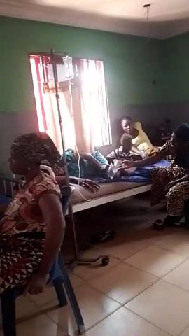 The video shows the interior of what appears to be a hospital room. There are several beds visible, some occupied by patients. One patient is connected to an IV drip. Several people are sitting in the room, including at least two who seem to be visiting or attending to patients. The room has tile floors and walls painted in a combination of green and grey. There is a window with red curtains. The video is shaky and the focus is unclear at times.