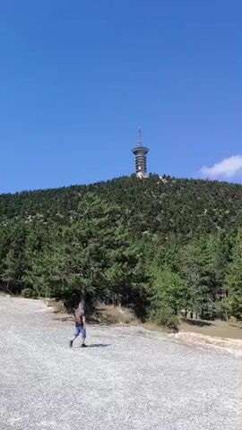 A person walks along a gravel path on a sunny day with a clear blue sky. In the background, a lush green forested mountain rises, topped by a tall, modern-looking tower with a circular structure at its peak. The path is bordered by trees and some rocky terrain. The camera pans to the right, revealing more trees and some yellow and red traffic cones. Further along, a wooden information kiosk with a roof is visible, also with traffic cones nearby. The path continues uphill, with more trees and rocky outcrops.