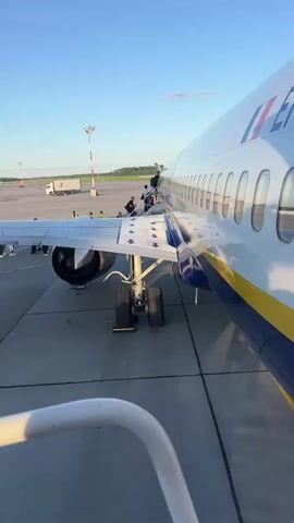 The video opens with a low-angle shot from the perspective of someone ascending a portable airstair, looking towards the right side of a parked airplane. The aircraft's landing gear, including its wheels and struts, is prominently visible. The fuselage of the airplane, painted white with a blue and yellow stripe along the bottom, stretches into the distance. Several passengers, dressed in casual clothing, are walking up the airstair and boarding the plane. Further in the background, other passengers can be seen on the tarmac. The scene takes place on an airport tarmac under a clear blue sky, with the sun setting, casting a warm glow on the surroundings. In the far distance, the outline of a cargo truck and airport infrastructure can be observed.