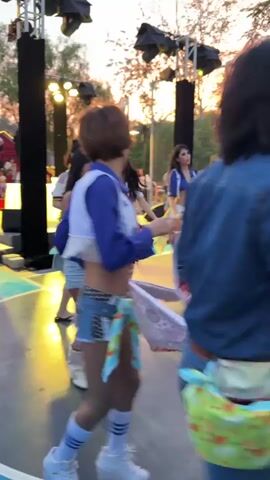 A group of people, including several women and children, are dancing on a paved outdoor area that appears to be a basketball court. The sun is setting, casting a warm glow and visible in the background. There are stage lights and speakers set up, suggesting some sort of performance or event. The women are dressed in casual, summery attire, with some wearing crop tops and shorts. The children are also dressed in casual clothing. Some of the people are holding and waving colorful fabric pieces, adding to the festive atmosphere. The overall scene depicts a lively outdoor gathering with music and dancing.