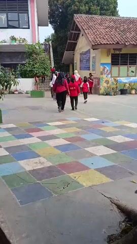 The video shows a group of children and adults in an outdoor schoolyard. The ground is paved with colorful squares, resembling a hopscotch court. To the left, a pink and white building with multiple windows is visible. To the right, a yellow building with a red tiled roof has murals painted on its exterior walls. Several children, mostly wearing red shirts and some in pink pants, are walking or standing in the schoolyard. Some adults, also in red shirts, are present. One adult is seen walking towards the left building, while others are gathered near the yellow building. The children appear to be engaged in various activities, some walking, others standing or looking around. The overall scene suggests a lively school environment during the daytime.