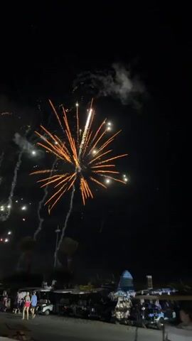 The video shows a nighttime fireworks display. Several bursts of fireworks, primarily in orange and red, illuminate the dark sky. Some fireworks leave trails of sparks as they descend. In the foreground, silhouettes of people are visible, some standing and others seated in what appear to be golf carts. A few palm trees are also discernible against the night sky. The overall scene suggests an outdoor celebration or event.