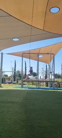 The video shows a woman riding a zipline swing in a playground. She is wearing a dark top, jean shorts, and loafers. The zipline is under a canopy held up by metal poles. The playground has a green artificial turf surface. In the background, there are other playground structures, trees, and a clear blue sky. The woman looks towards the camera and smiles during the ride.
