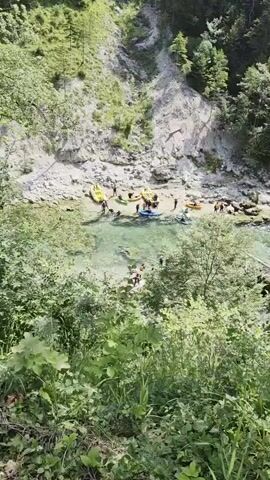 A group of people are gathered at the edge of a clear, shallow river. Several inflatable rafts and kayaks are visible on the shore and in the water. Some individuals are standing in the water, while others are preparing to get into the watercraft. The river flows through a mountainous area with steep, rocky banks covered in dense green trees and vegetation. The sunlight is bright, suggesting a clear day.