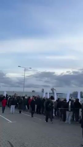 The video shows a large group of people standing behind a barrier on a paved area, watching something in the distance. The crowd consists of people of various ages and wearing casual clothing. In the background, there are several white flags with lettering and logos, as well as tents, trees, and possibly buildings. The sky is overcast with a mix of clouds and patches of blue. The camera pans from left to right, revealing more of the crowd and the background. There are street lights scattered throughout the area. In the middle of the video, a white car is briefly visible performing a stunt. The overall scene appears to be an outdoor event or gathering.