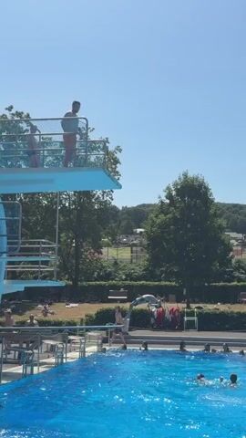 This video captures a bright, sunny day at an outdoor swimming pool in Bernkastel-Kues, Germany. The scene is set with a clear blue sky and lush green trees in the background, suggesting warm weather. In the foreground, the large pool is filled with vibrant blue water, dotted with several people swimming and enjoying the day.

The main action focuses on a person who has just leaped from a high diving board. They are captured mid-air, performing a flip or acrobatic maneuver before plunging into the water, creating a significant splash. The diving board itself is elevated and has safety railings. Below the diving board, other swimmers are visible, some are near the splash zone while others are further away.

In the background, there are benches, more trees, and a structure that appears to be a lifeguard station or changing area. A red and white windsock is visible atop this structure. Several people are gathered on the grass and around the pool, seemingly relaxing or spectating. The presence of other swimmers and the overall atmosphere suggest a recreational and leisurely event. The clear daylight indicates it is daytime, likely mid-morning to late afternoon. There are no discernible emotions other than general enjoyment of a summer day at the pool. No visible text is present in the video.