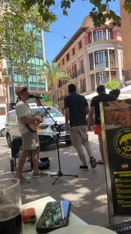 A man wearing a straw hat and red band is playing a ukulele and singing into a microphone on a cobblestone street. He is wearing a white t-shirt and light-colored shorts. Next to him is a small black amplifier and speaker. A white car is parked on the street behind him. In the background, there are buildings with balconies and awnings, as well as trees and other pedestrians. In the foreground, there is a table with a plate, fork, glass of dark liquid, and a smartphone. A yellow sign advertising hamburgers is visible on the right. The scene appears to be a sunny day in a European city.