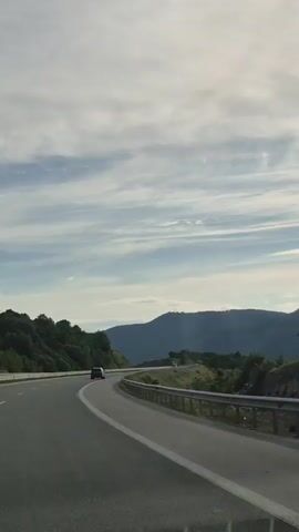 The video shows a car driving on a highway. The sky is cloudy with patches of blue and the sun is visible. There are mountains and trees in the background. The road is paved and has white lines to mark the lanes. There are guardrails along the side of the road. A few other cars can be seen on the highway in the distance. The video appears to have been filmed from inside a moving vehicle.