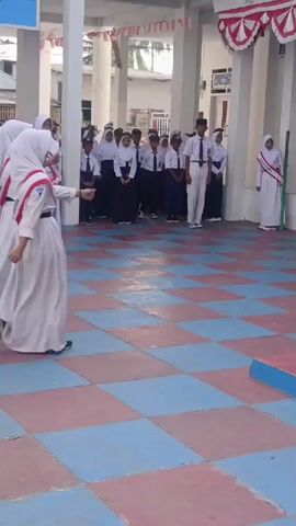 Three young women in white uniforms and headscarves are marching in formation on a checkered floor. They are decorated with red and white sashes. The background shows a building with white columns and red and white decorations. Other students are standing in the background watching.