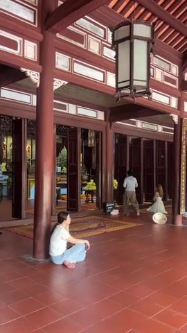 A wide shot shows the interior of a large, traditional building with a red tiled floor and ornate wooden architecture. In the foreground, a young woman in a white t-shirt and light blue pants sits on the floor with her legs crossed, facing away from the camera. She is leaning against a thick red pillar. In the background, several other people are visible. To the right, a woman in a white dress stands near a doorway. Further back, a person in light-colored clothing is also standing. The building features intricate carvings, large wooden doors, and hanging lanterns. The overall atmosphere appears serene and spiritual, possibly a temple or pagoda.