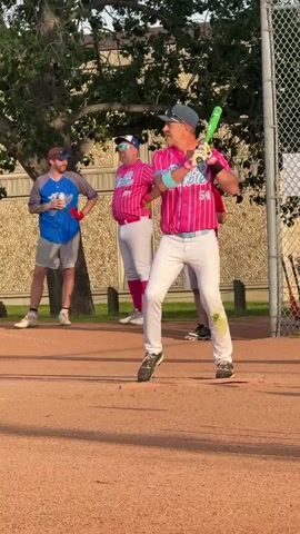 A man in a pink and blue striped baseball jersey with white pants and a baseball cap is holding a green baseball bat and appears to be in a batting stance on a baseball field. He is looking towards the left of the frame. Two other men are standing in the background. One man on the left is wearing a blue shirt and gray shorts and is holding a drink. Another man in the center is wearing a pink and blue striped jersey and white pants. A chain-link fence is visible on the right side of the frame. The ground is covered in reddish-brown dirt. A tree is on the left side of the frame.